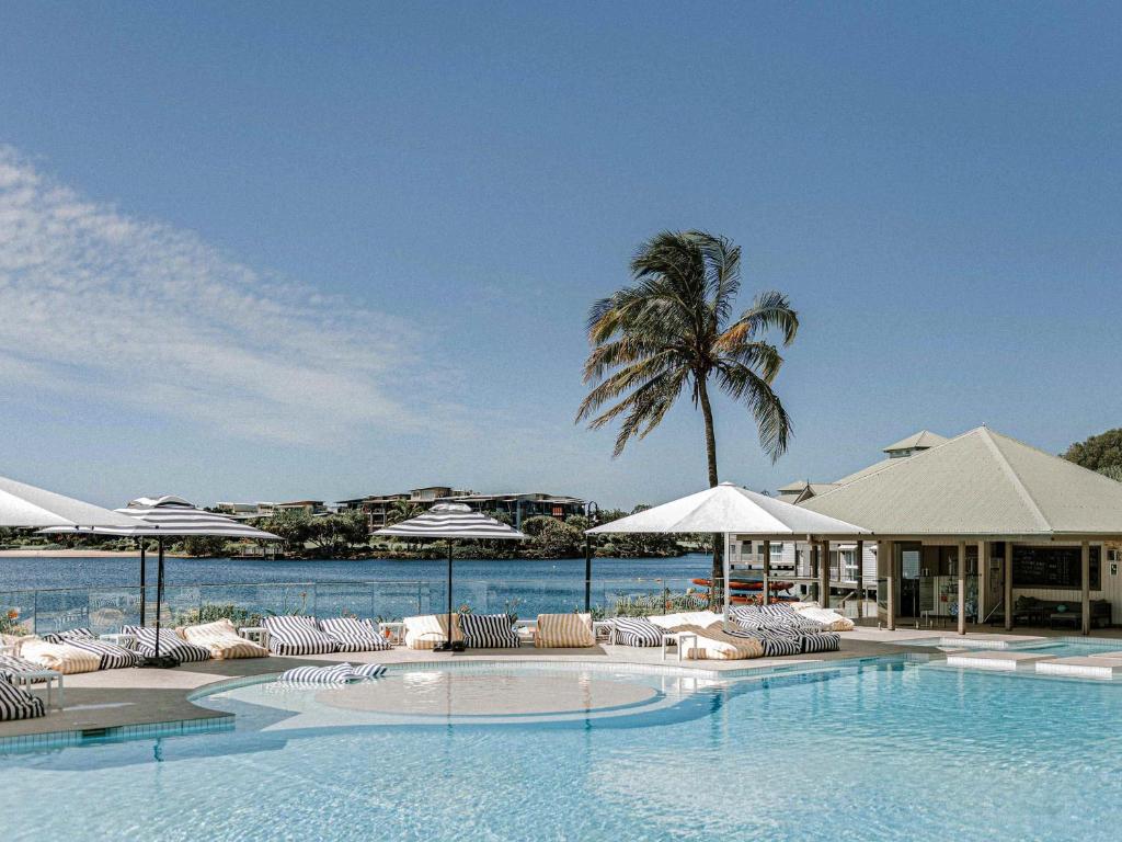 a swimming pool with chairs and umbrellas and a palm tree at Novotel Sunshine Coast Resort in Twin Waters
