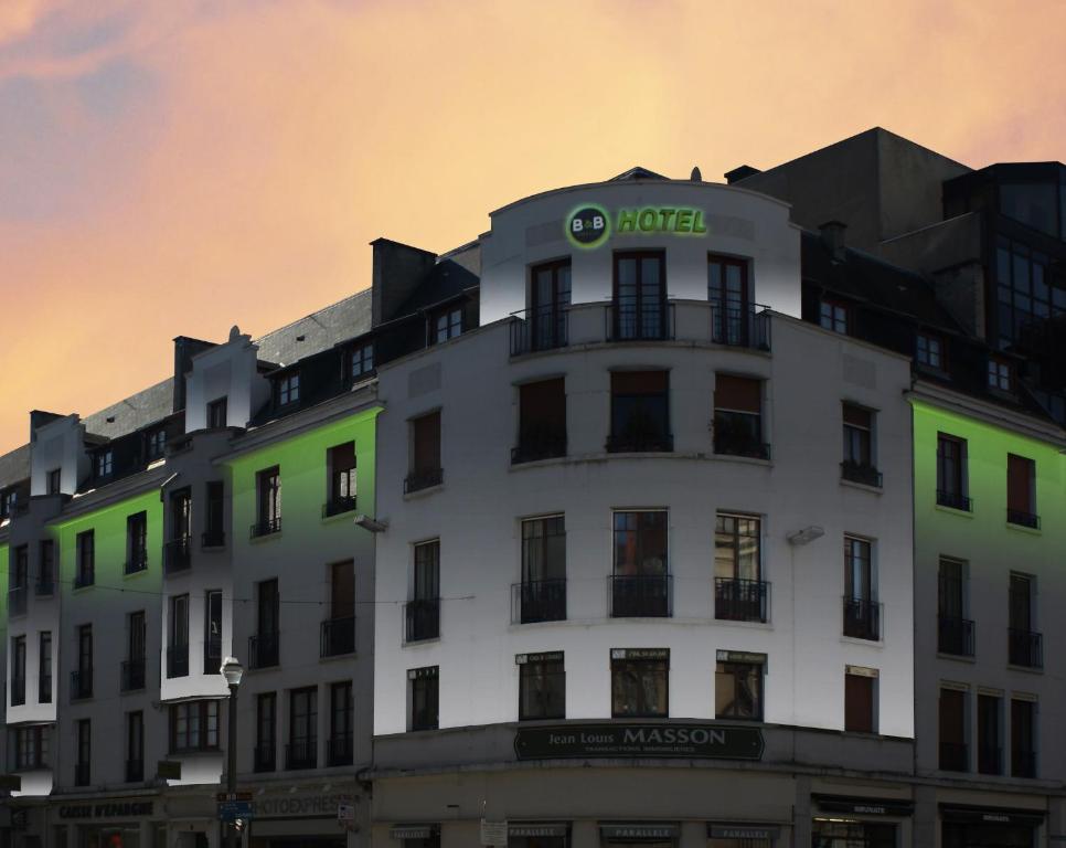 Prédio de esquina de vários andares ao entardecer, com fachada branca e letreiro iluminado “B&B HOTEL” no topo, janelas com sacadas e luzes verdes destacando partes da construção, para representar hotéis em Dijon.