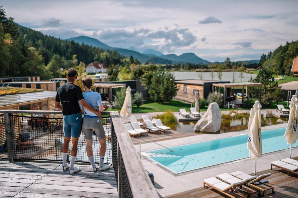 a couple standing on the balcony of a hotel with a pool at Lakeside Petzen Glamping in Unterlibitsch