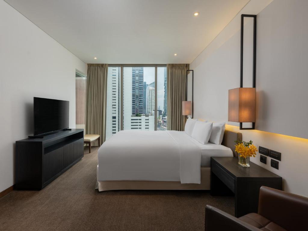 A bright guest room centers a large bed opposite a television and city-facing window.