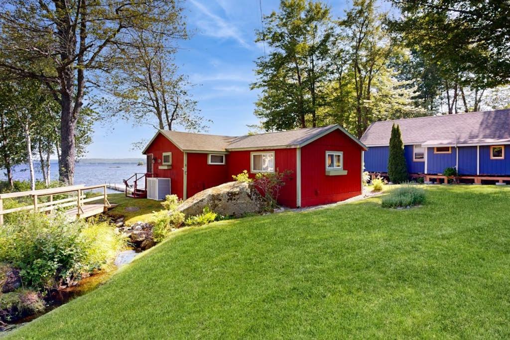a red house on a lawn next to the water at Secluded lakefront cottage great views, dock & boat launch in Ellsworth