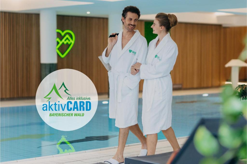 a man and woman in white robes standing next to a swimming pool at BERGlässig Hotel - Halbpension Plus inkl Getränke in Bodenmais