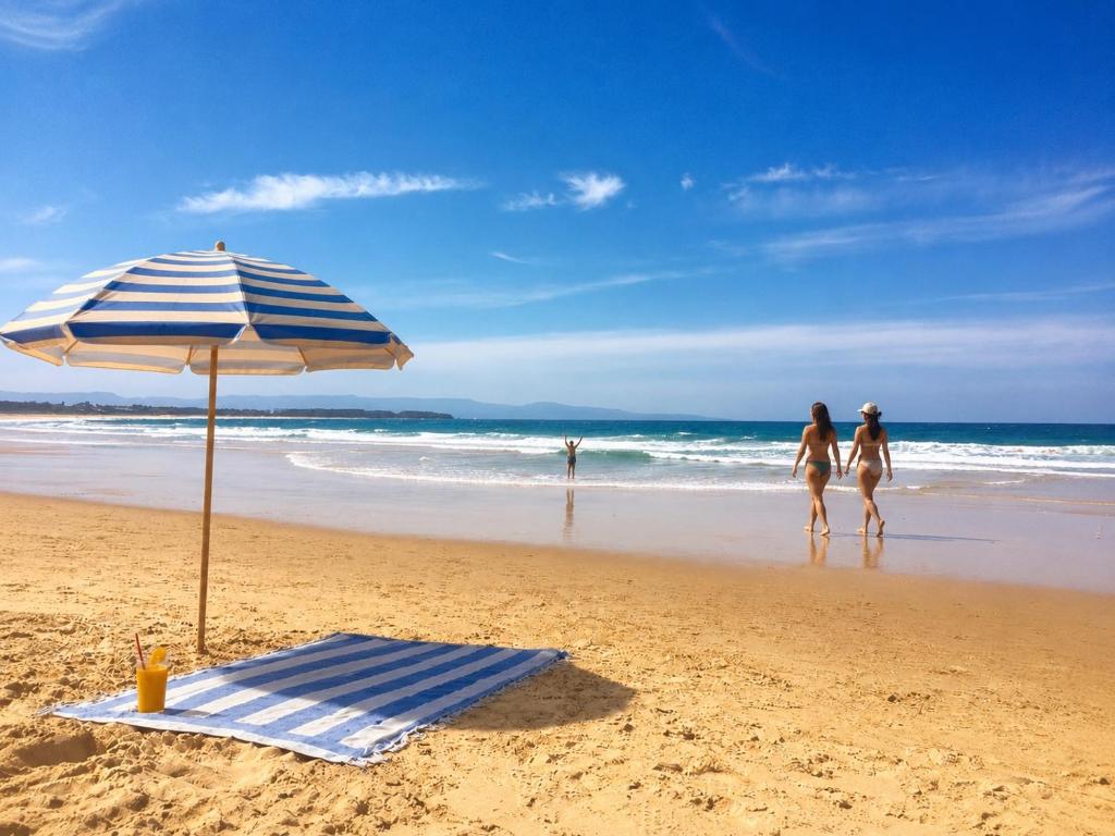 two women walking on the beach with an umbrella and a towel at Palacete Beach - Resort Style Stay in Culburra Beach