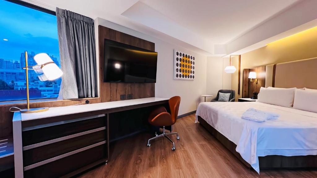 A room with a bed, wall-mounted television, desk and city-facing window.