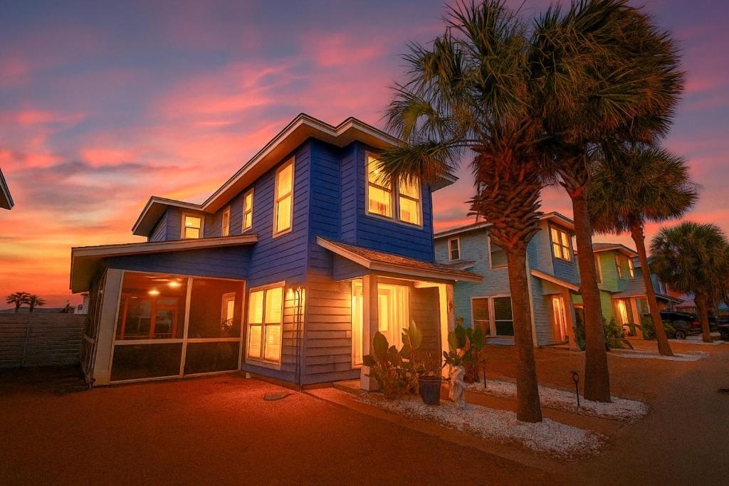 a blue house with palm trees in front of a sunset at Salty Waves | Beach Cottage, Walk to Beach, Pool, Fenced Yard & Grill in Port Aransas