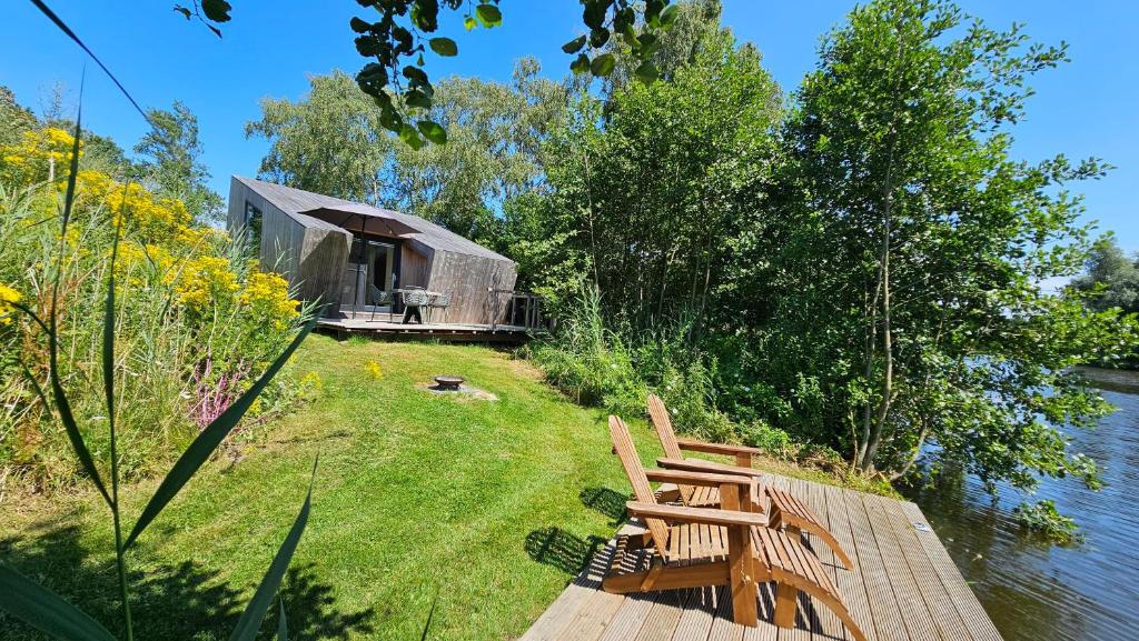 a cabin on the side of a lake with two wooden chairs at Tiny House De Klyster in Westergeest