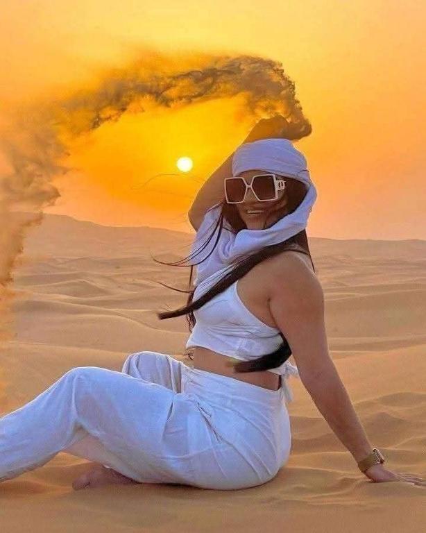 a woman sitting in the desert with the sun in the background at Fayoum Safari Camp in Qaryat at Ta‘mīr as Siyāḩīyah