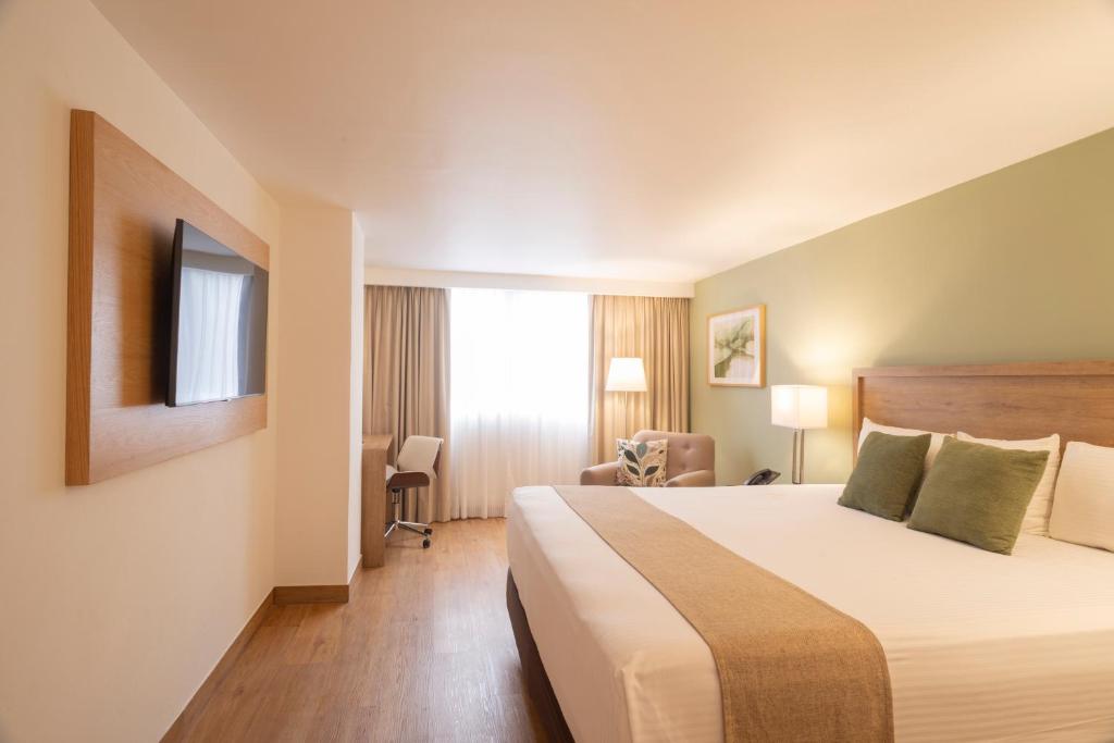 A wide bedroom view with the bed, television, window, desk and armchair in one frame.