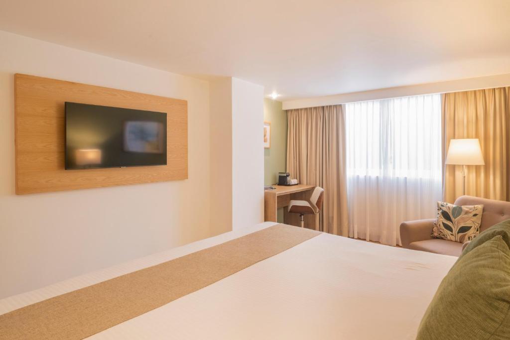 A bedroom view showing the bed, wall-mounted television, window, desk and armchair.