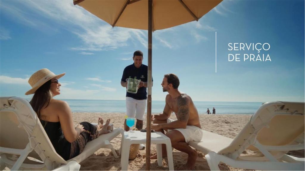 three people sitting on the beach under an umbrella at Hardman Praia Hotel in João Pessoa