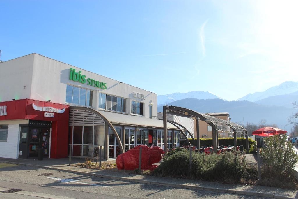 a building with a store with mountains in the background at Ibis Styles Crolles Grenoble A41 in Crolles