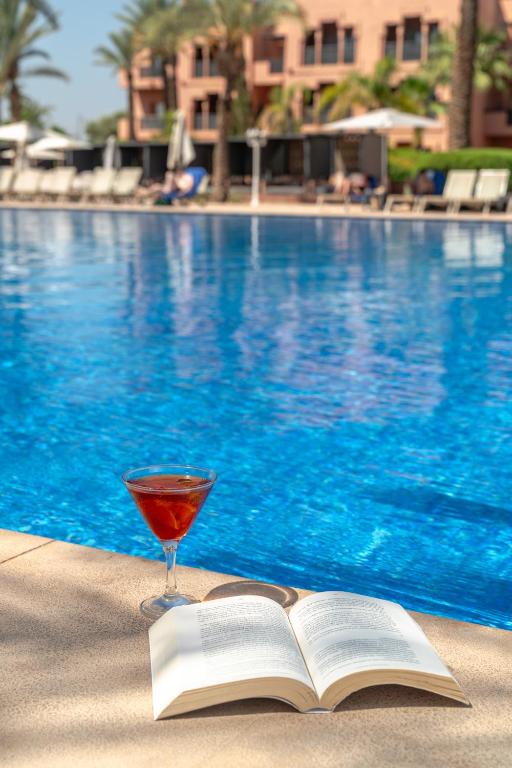 a book and a glass of wine next to a swimming pool at Kenzi Menara Palace & Resort in Marrakesh