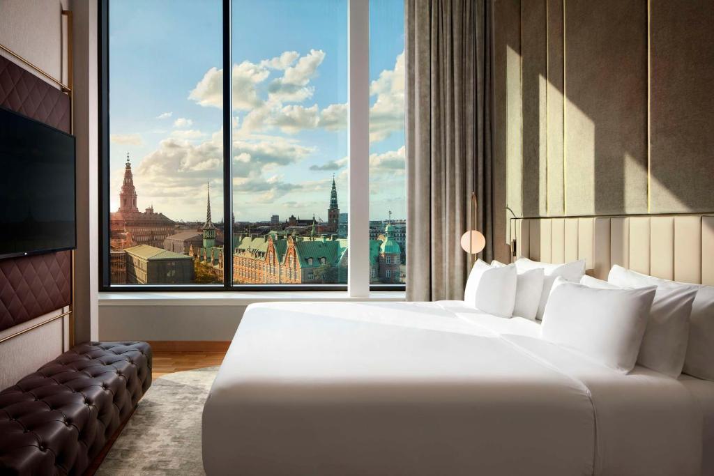 Quarto do NH Collection Copenhagen, Copenhague, com cama grande de casal e lençóis brancos, uma TV de frente para a cama, com um sofá marrom logo abaixo dela e uma grande janela, com vista para pontos turísticos da cidade, para representar Dinamarca.