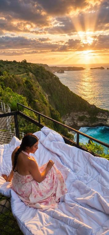 a woman sitting on a bed on a balcony with the ocean at The Indah Cliff Sea View in Polilit