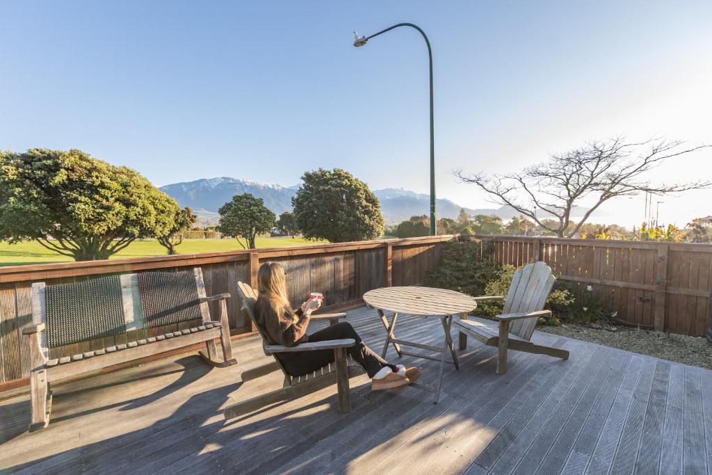 a woman sitting on a chair on a deck with a table at Handy Location, Ocean & Mountain Views, Close to Park - KK96845 in Kaikoura