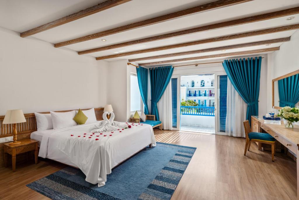 A bright bedroom with a large white bed, blue curtains, timber ceiling beams and an open glass doorway.