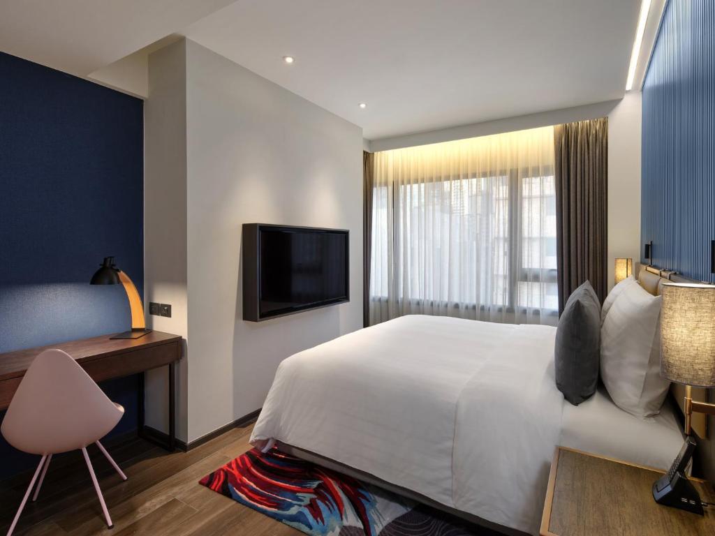 A bedroom includes a narrow writing desk beside a large bed and a wall-mounted television.