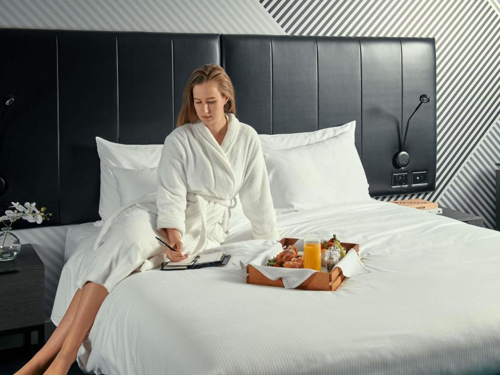 a woman sitting on a bed with a tray of food at Movenpick Hotel Melbourne On Spencer in Melbourne