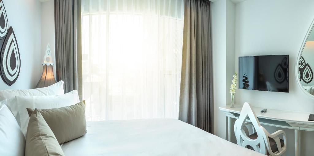 A bright bedroom with a white bed, large curtained window, television and compact desk.