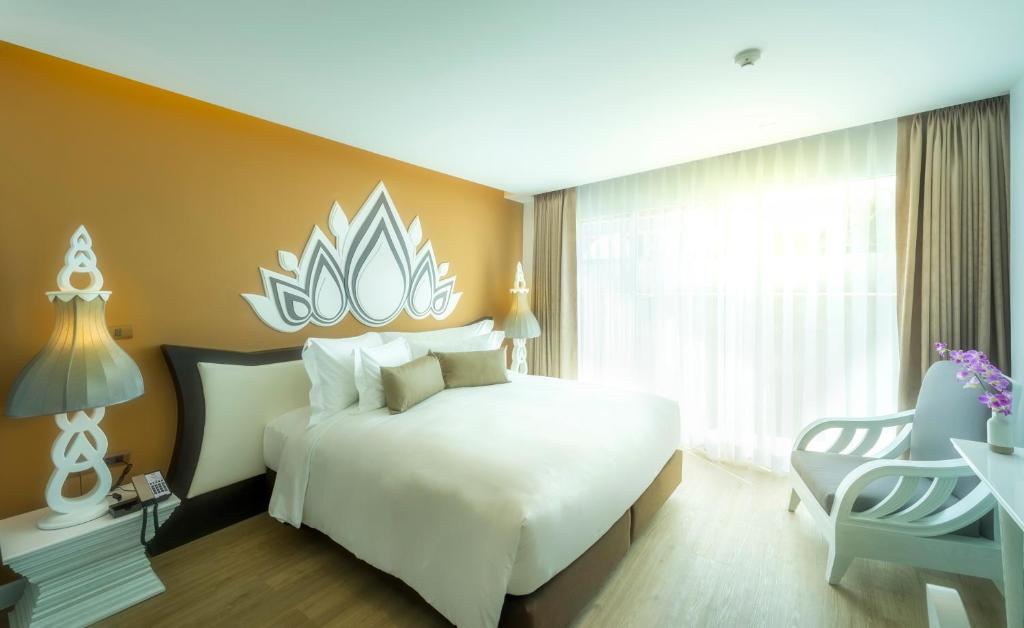 A warm-toned bedroom with a large bed, ornate Thai-style headboard, and two chairs by the window.