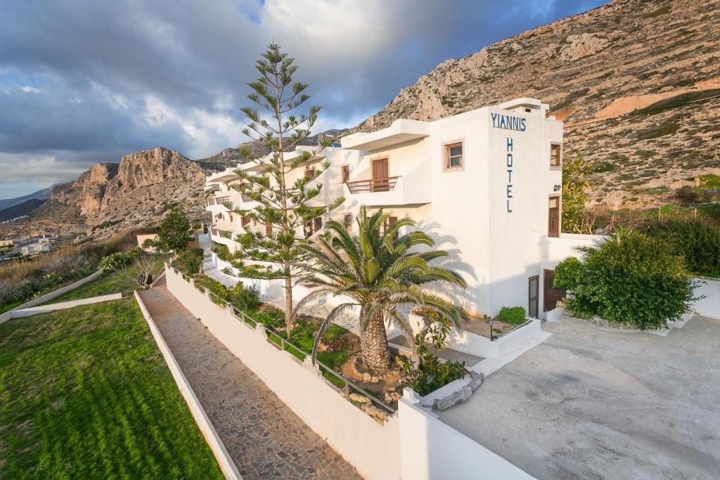 a white building with palm trees in front of a mountain at Yiannis Ηotel in Arkasa