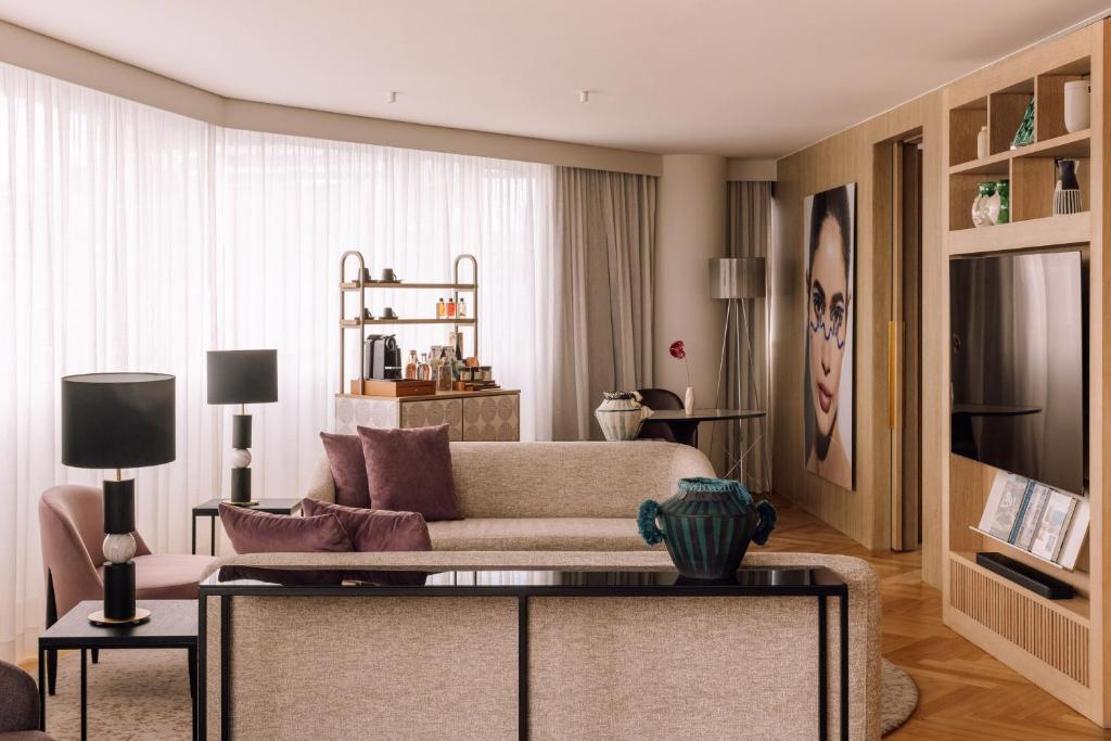A bright lounge facing a broad curtained window, with sofa seating, television and drinks station.
