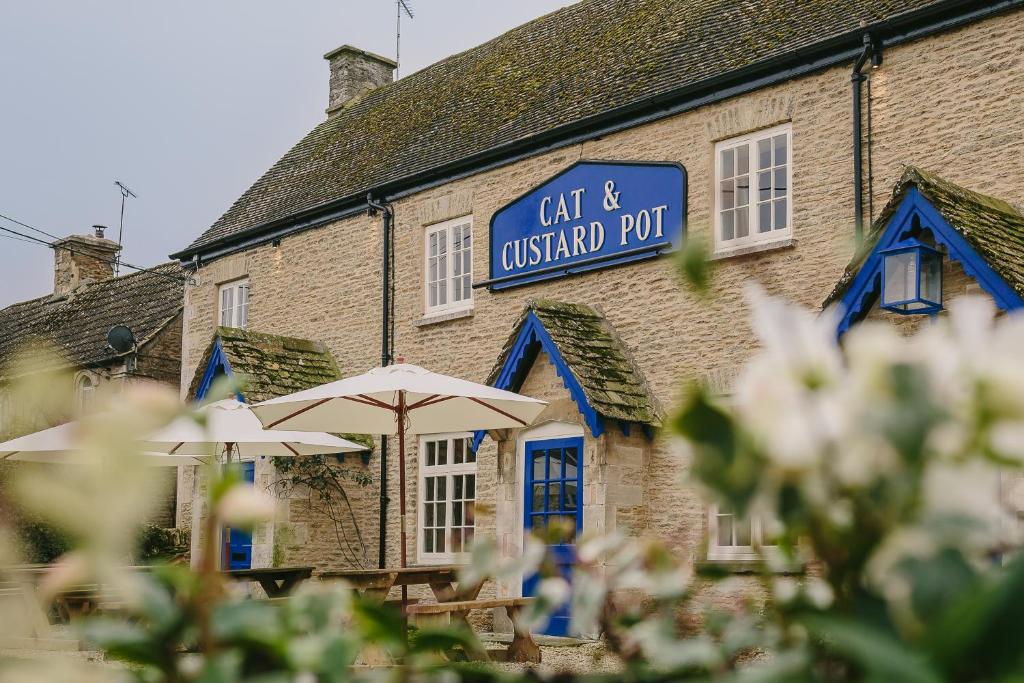 un edificio con sombrillas y un cartel en él en Cat and Custard Pot Inn en Shipton Moyne