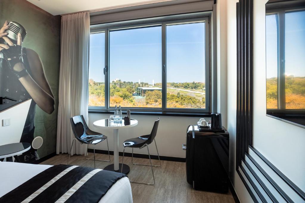 A bright corner room with a round table, two chairs and broad windows overlooking greenery and city buildings.