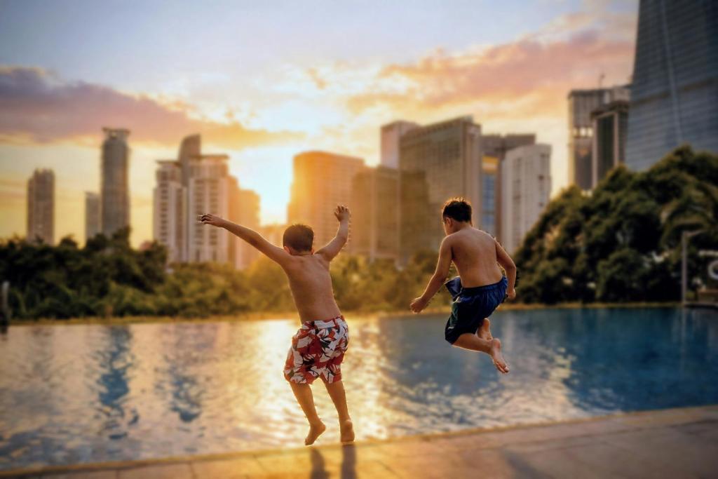 Two children jump into a rooftop-looking pool against a city skyline at sunset.