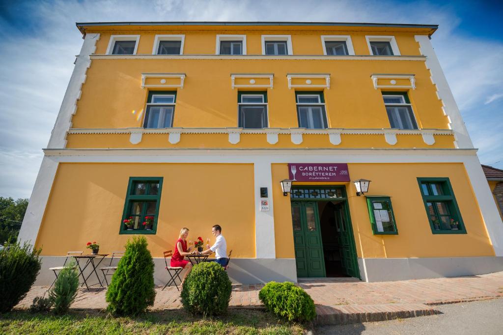 una pareja de pie en frente de un edificio amarillo en Hotel Cabernet, en Villánykövesd