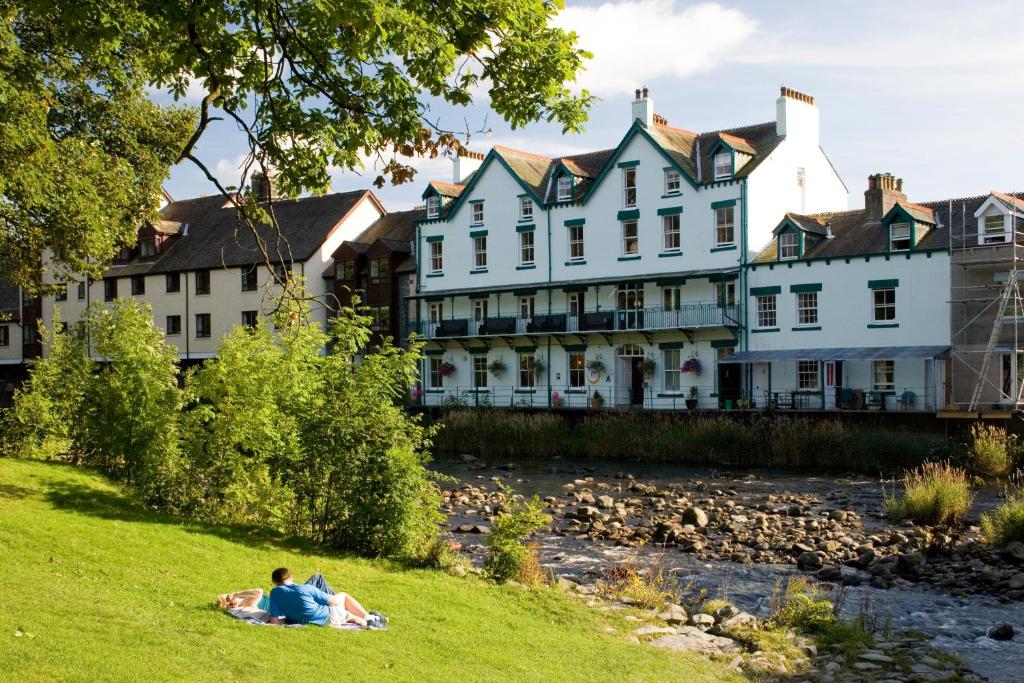 two people laying on the grass next to a river with buildings at YHA Keswick in Keswick