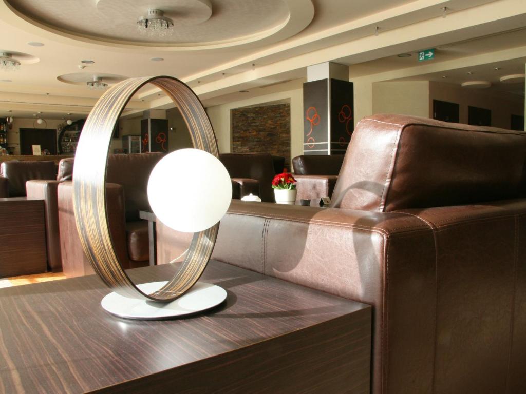 Contemporary circular table lamp on dark wood side table beside brown leather sofas, sunlit lobby.
