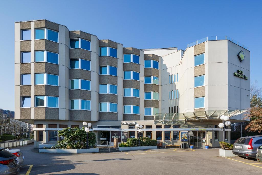 een groot gebouw met auto's geparkeerd op een parkeerplaats bij Hotel Welcome Inn in Kloten
