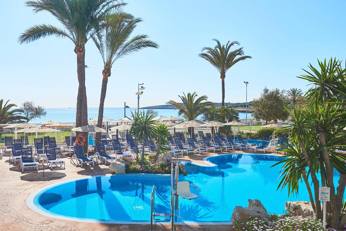Una piscina con sillas y palmeras y el océano. en Hipotels Hipocampo Playa, en Cala Millor