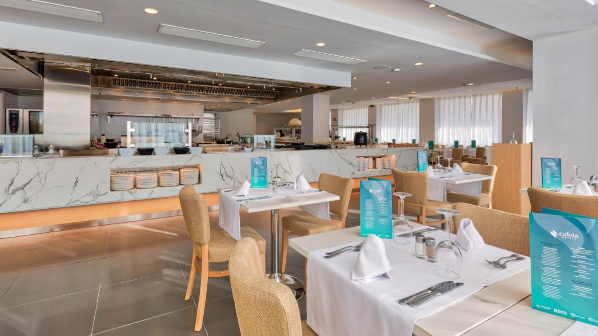 Un restaurante con mesas y sillas blancas y una cocina. en Talayot - Adults Only, en Cala Millor
