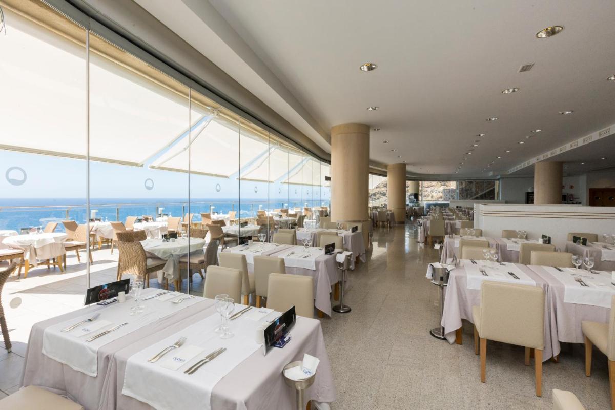 Un restaurante con mesas y sillas blancas y grandes ventanales. en Gloria Palace Amadores Thalasso & Hotel, en Puerto Rico de Gran Canaria