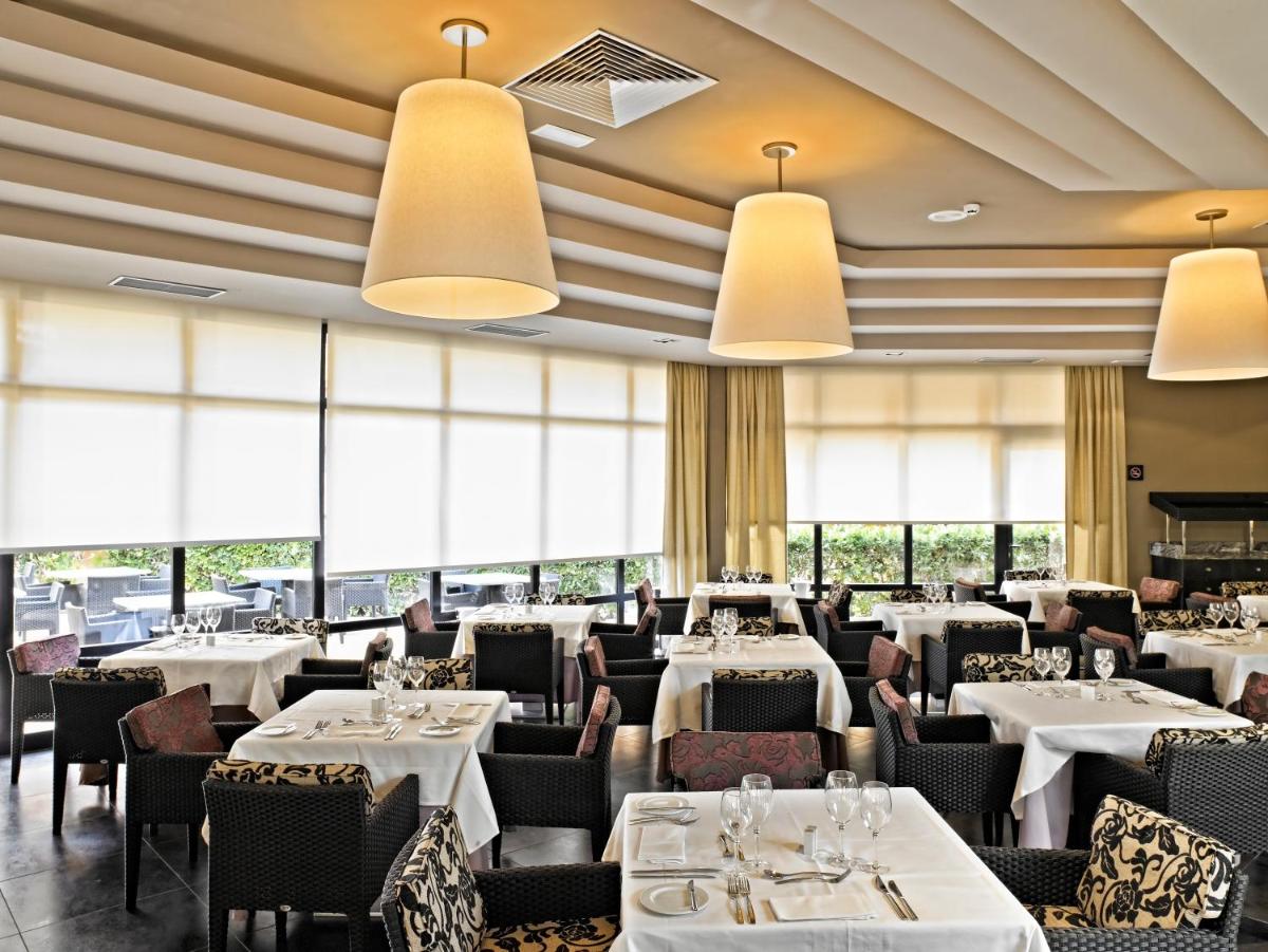 Un restaurante con mesas y sillas blancas y grandes ventanales. en Protur Turo Pins 4* SUP, en Cala Ratjada