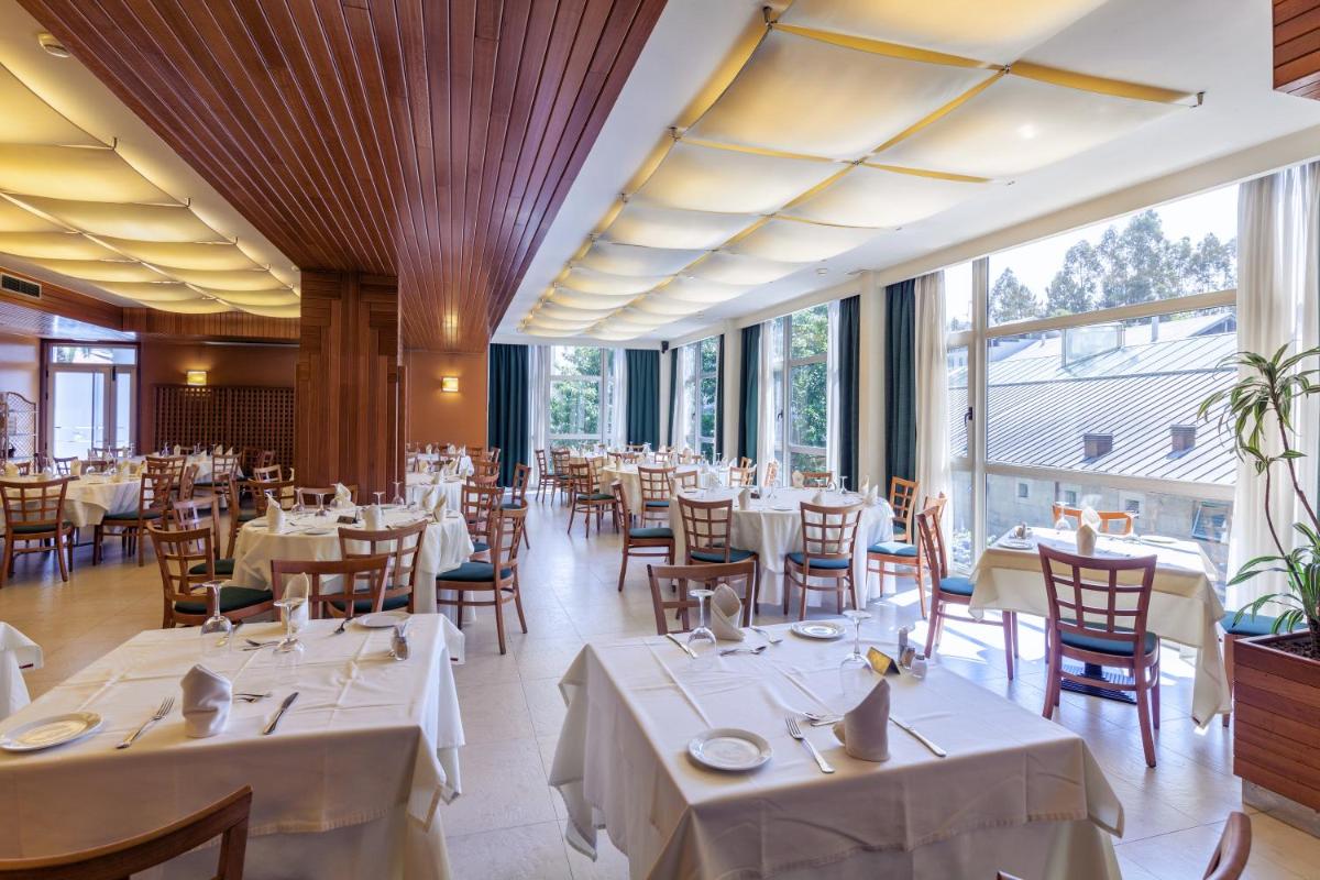 Un restaurante con mesas y sillas blancas y grandes ventanales. en Hotel Balneario de Compostela, en Brión