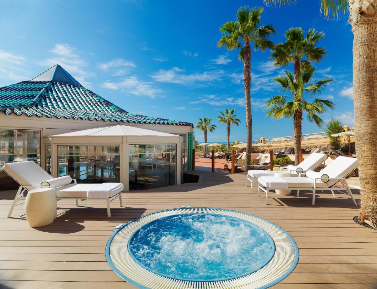 Una terraza con piscina, sillas y palmeras. en H10 Playa Esmeralda - Adults Only, en Costa Calma