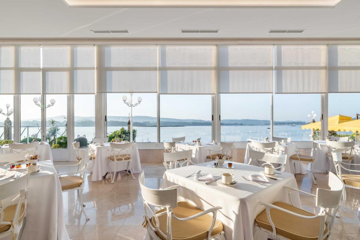 Un restaurante con mesas y sillas blancas y grandes ventanales. en Eurostars Gran Hotel La Toja, en Isla de la Toja