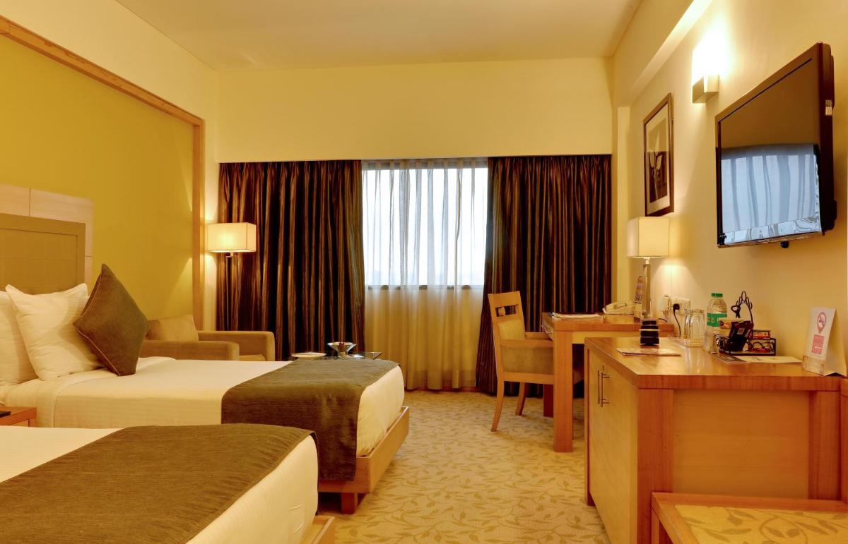a hotel room with two beds and a desk at The Suryaa New Delhi in New Delhi a hotel room with two beds and a desk at The Suryaa New Delhi in New Delhi