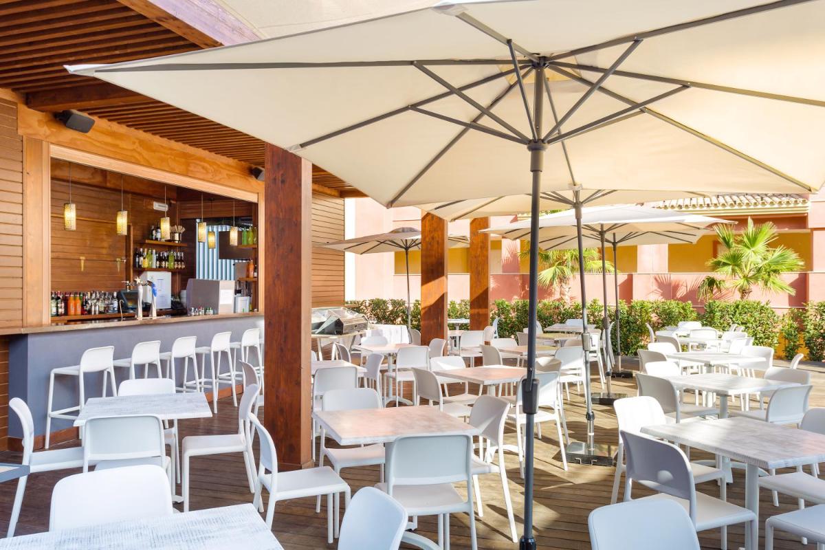 Un restaurante con mesas y sillas blancas y sombrillas. en Ama Islantilla Resort, en Islantilla