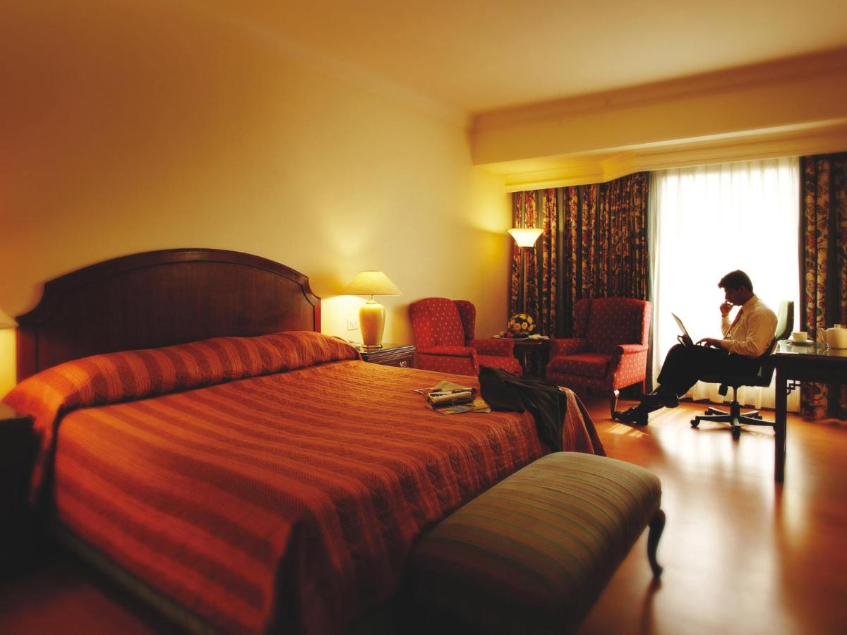 a hotel room with a bed and a man working on a laptop at The Lalit Mumbai in Mumbai