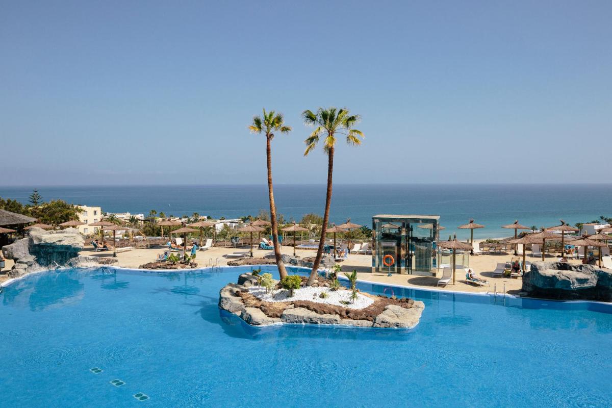 Una gran piscina con palmeras y el océano. en Alua Village Fuerteventura - All Inclusive, en Playa de Jandía