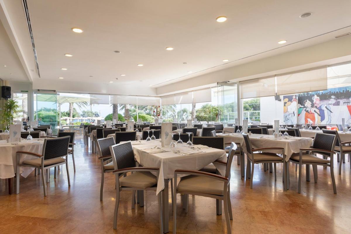 Un restaurante con mesas y sillas blancas y ventanas. en Hipotels Dunas Aparthotel, en Cala Millor