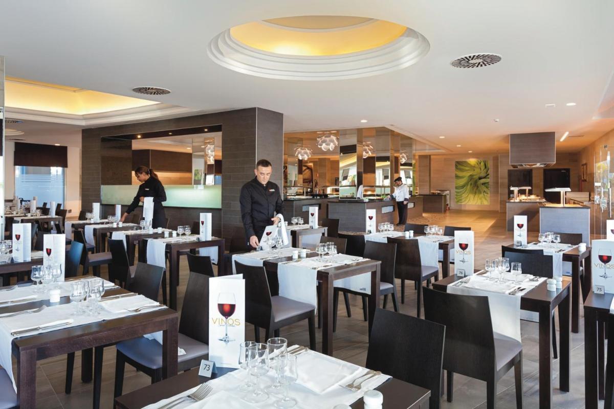 Un restaurante con mesas blancas y sillas negras y gente. en Hotel Riu Arecas - Adults Only, en Adeje