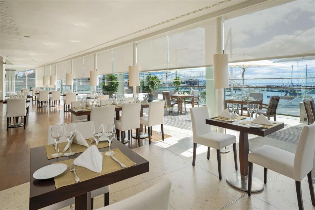 um restaurante com cadeiras brancas, mesas e janelas em Hotel Marina Atlântico em Ponta Delgada