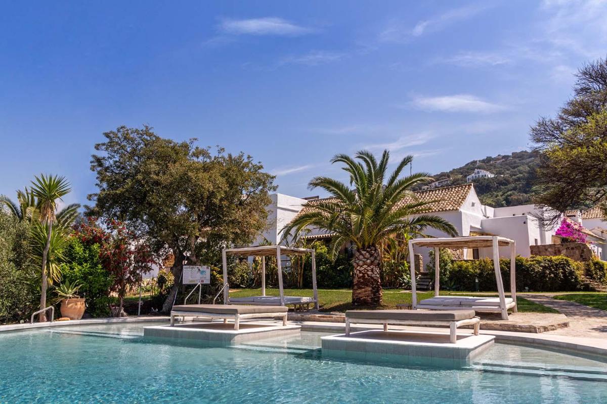 una piscina con bancos y árboles y un edificio en El Cortijo de Zahara by QHotels, en Zahara de los Atunes