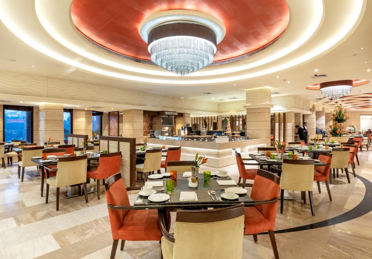 a restaurant with tables and chairs and a large ceiling at The Suryaa New Delhi in New Delhi a restaurant with tables and chairs and a large ceiling at The Suryaa New Delhi in New Delhi