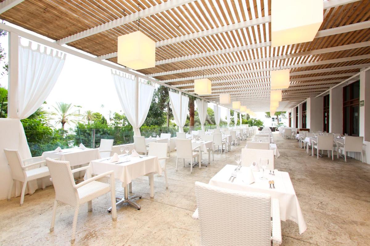 Un restaurante con mesas y sillas blancas y ventanas. en Sentido Fido Tucan - Adults Only, en Cala d´Or